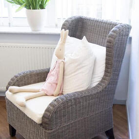 Geflochtener Sessel mit Kissen und Stoffhase im hellen Kinderzimmer vor Fenster.